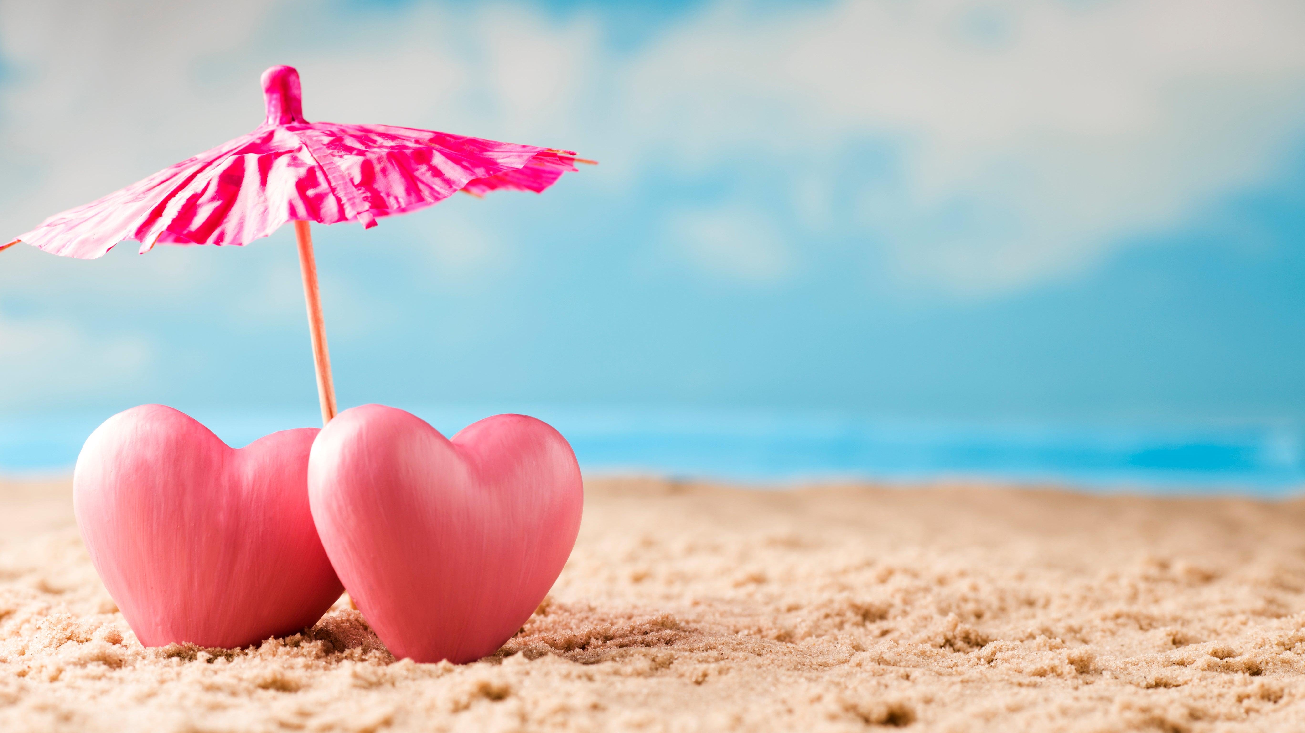 Two pink hearts on sandy beach under pink cocktail umbrella, blue sky in background