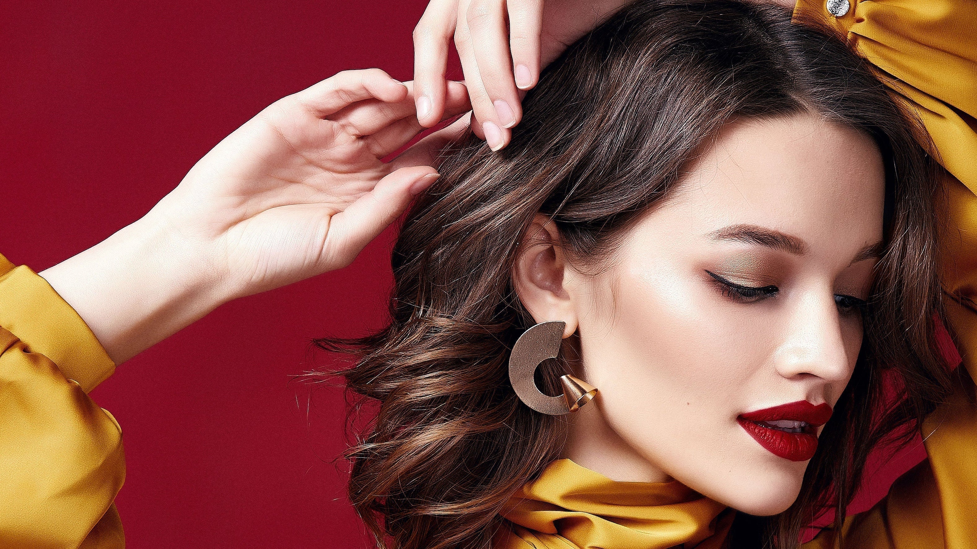 Woman with flawless makeup, bold red lipstick, gold eyeshadow, and large statement earrings, wearing a satin yellow blouse against a deep red background