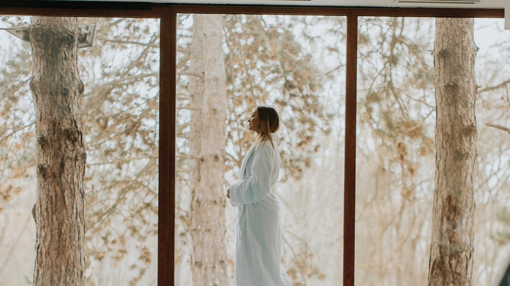 Woman in white robe standing indoors by window overlooking winter forest landscape