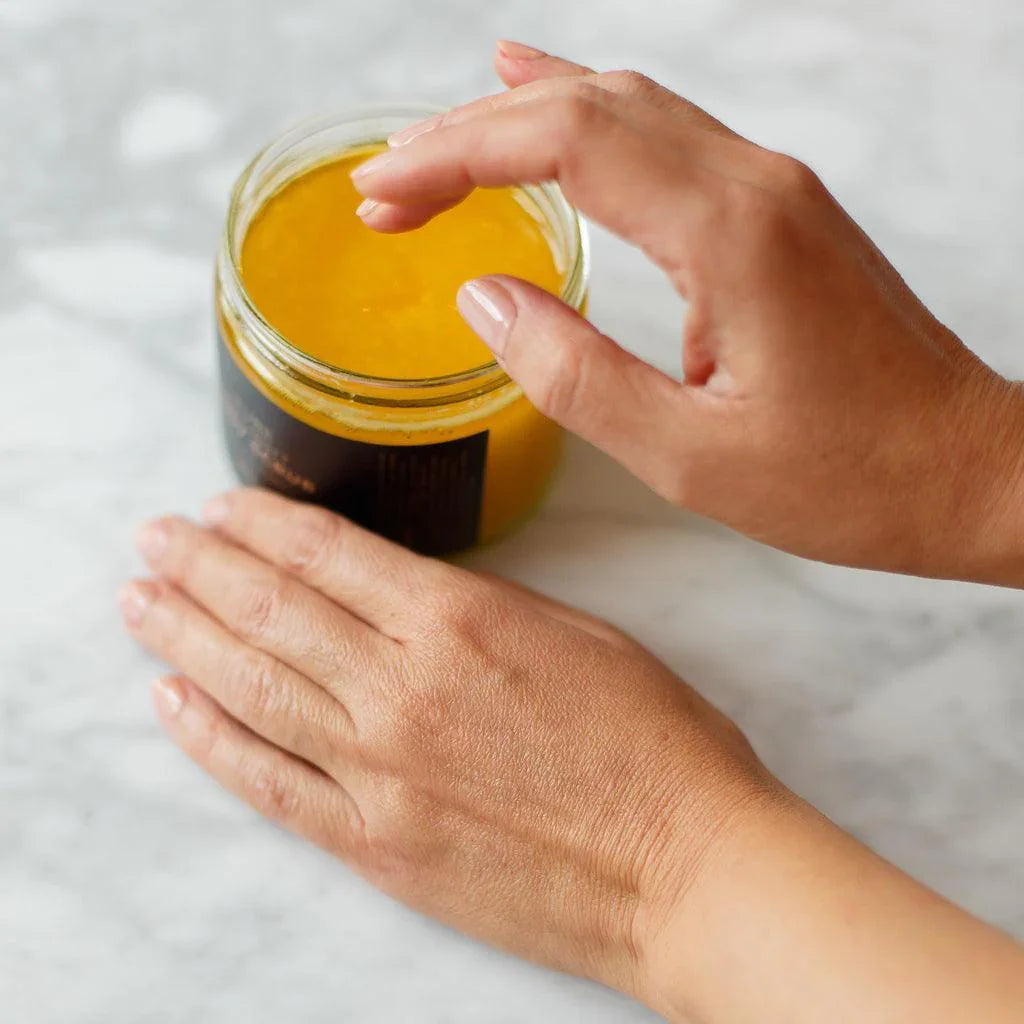 Hand scooping yellow body scrub from glass jar on marble surface