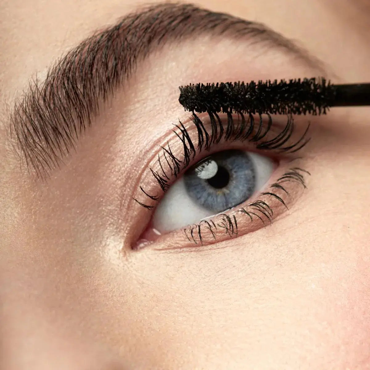 Close-up of blue eye with thick lashes applying black mascara wand