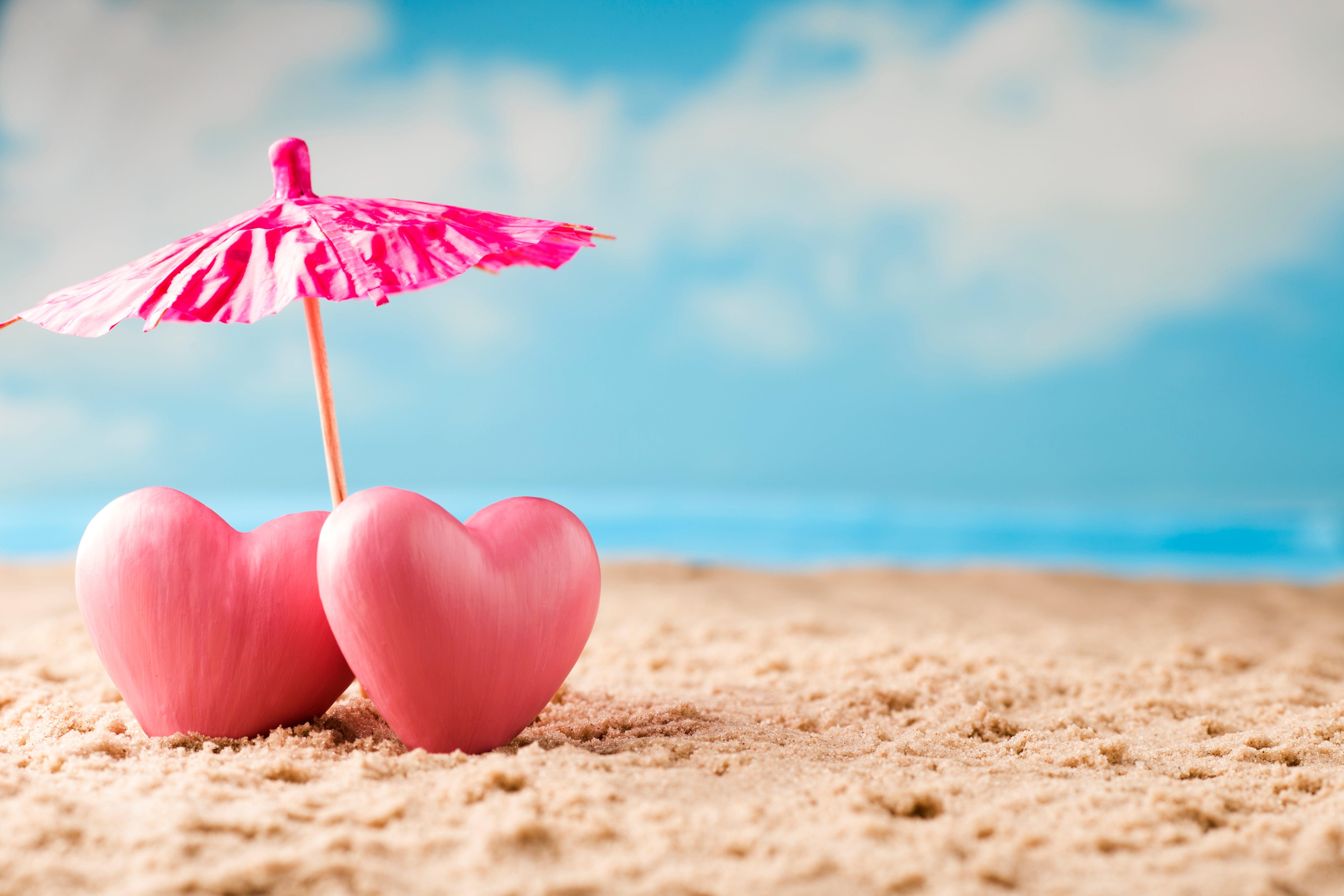 Two pink hearts on sandy beach under pink cocktail umbrella, blue sky in background