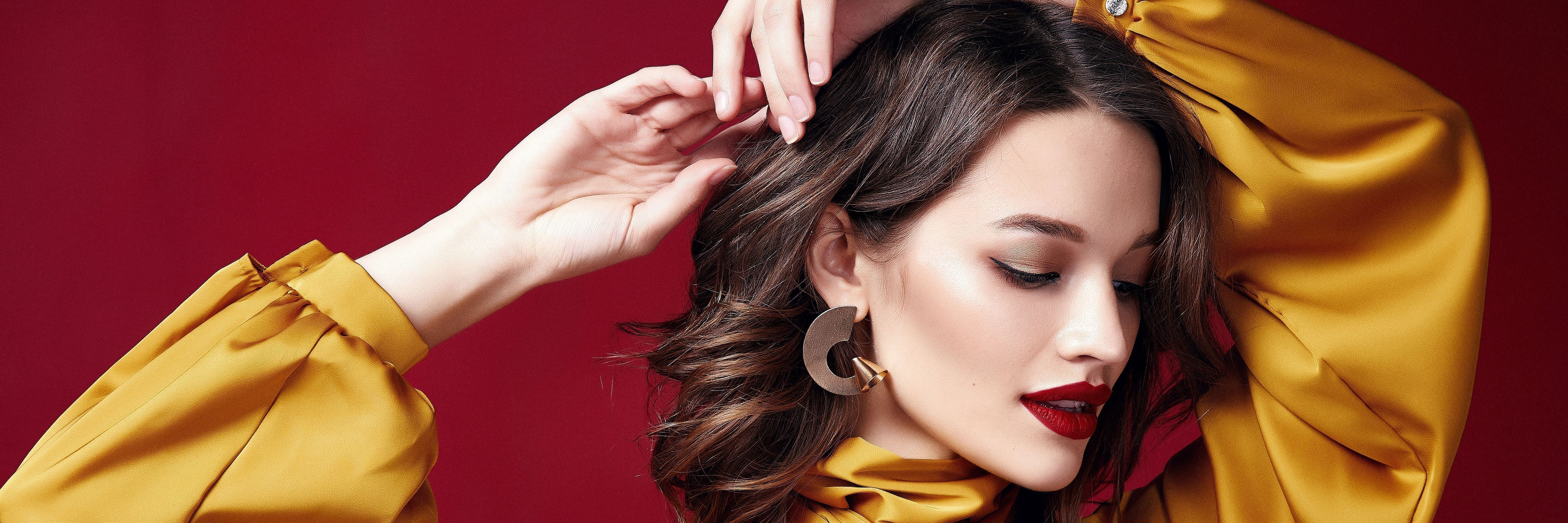 Woman with flawless makeup, bold red lipstick, gold eyeshadow, and large statement earrings, wearing a satin yellow blouse against a deep red background