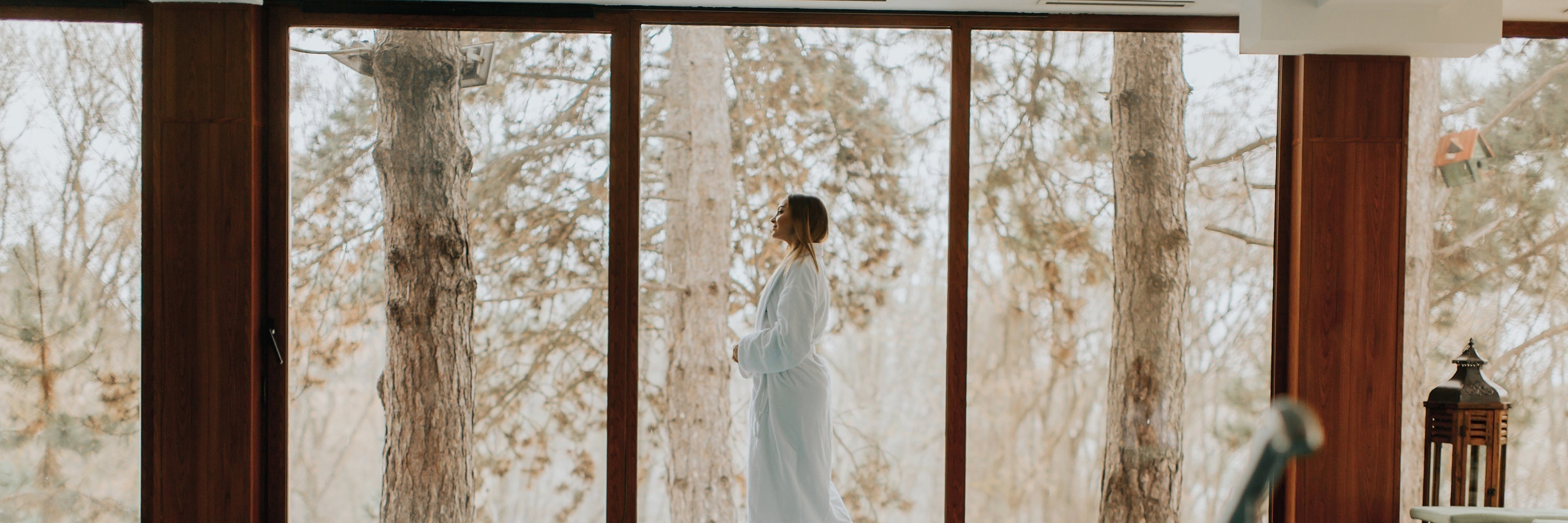Woman in white robe standing indoors by window overlooking winter forest landscape